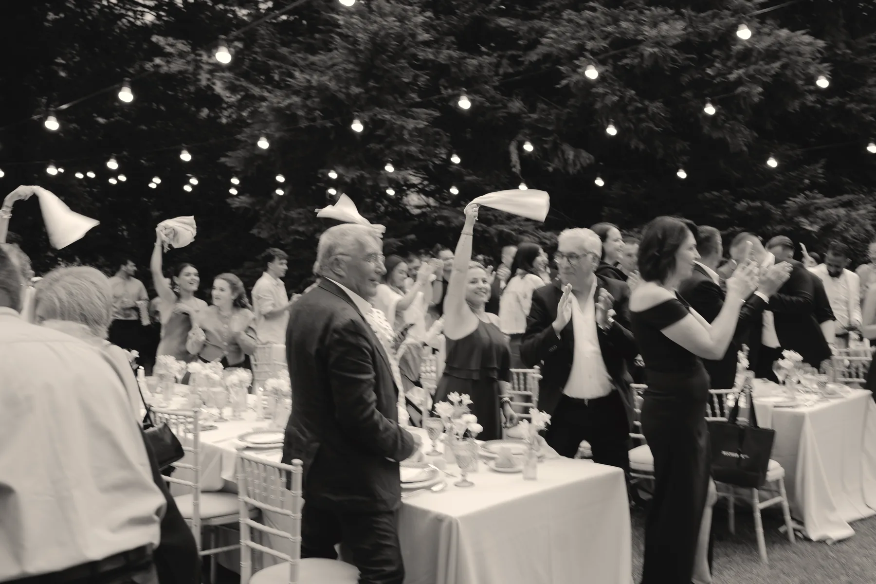 Les invités debout agitent leurs serviettes sous les guirlandes lumineuses, en noir et blanc